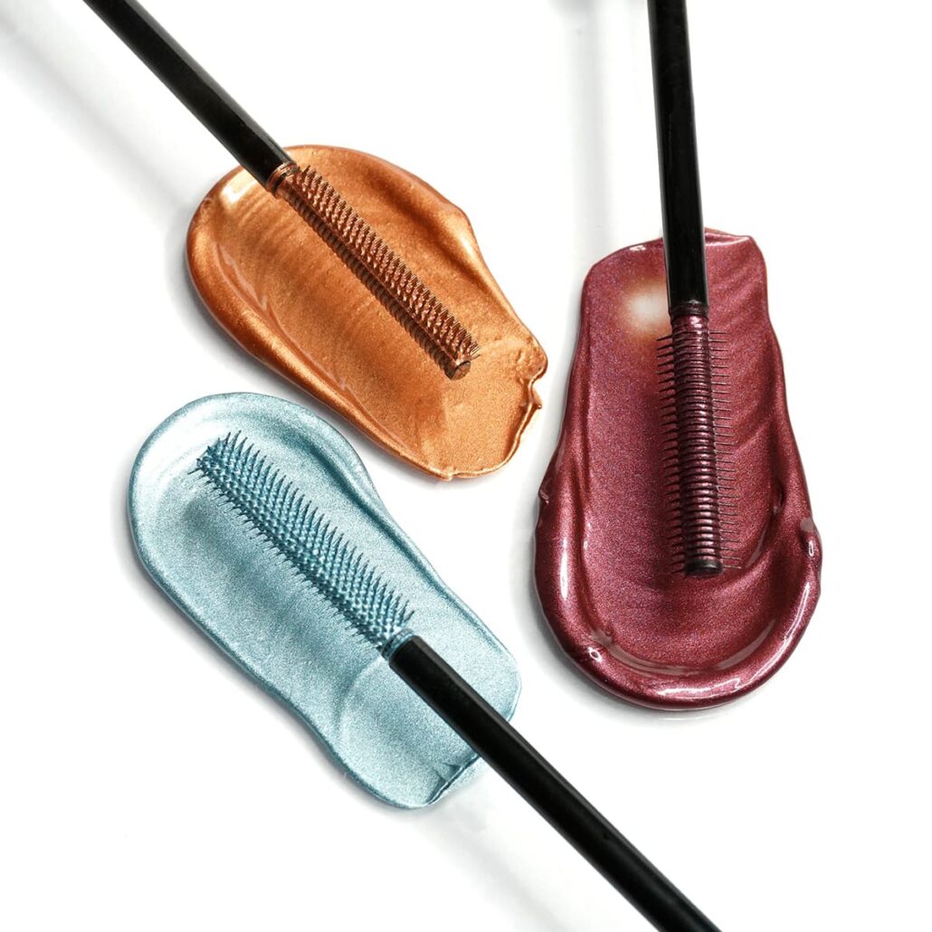 Three mascara brushes over metallic blue, bronze, and burgundy swatches